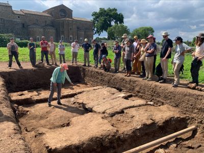 Archaeologist stands in a dig-site surveying the remains of a structure, while others stand around the site, looking on. The site is a green field with an old building in the distance.