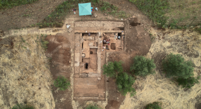 Overhead view of the Falerii Novi excavation site, showing people working to uncover sections of an ancient structure, with a blue tent nearby, amid some trees and grassland.