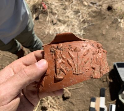 Archaeologist hand holding up a section of red ceramic showing dancing figures and symbols.
