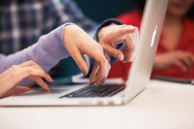 Close-up of multiple hands typing and pointing at a laptop keyboard, with people collaborating around the device