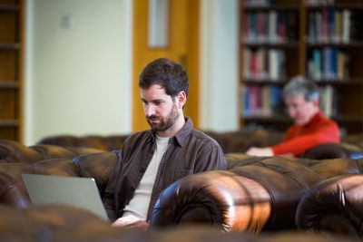Man using a laptop while seated on a brown leather sofa in a library, with another person reading in the background